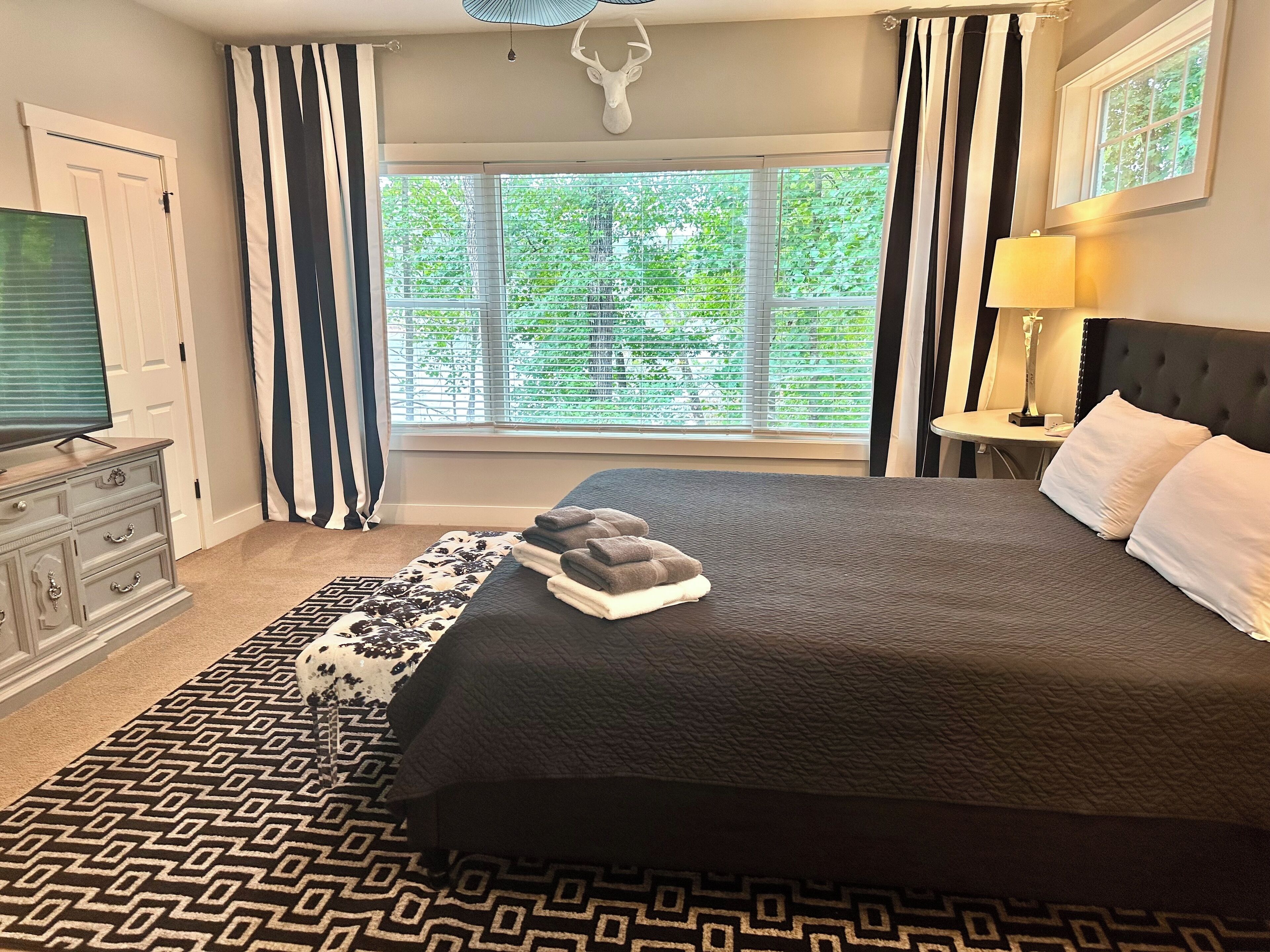 A bedroom with a large bed, a dresser, a window with blinds, a lamp, and a ceiling light.