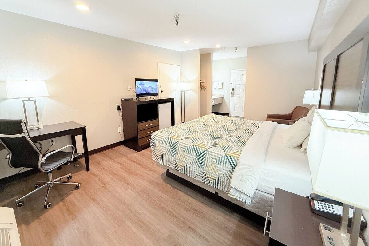 Une chambre d’hôtel avec un lit, un bureau avec un ordinateur, une chaise, une télévision et une salle de bain visible à travers une porte ouverte.