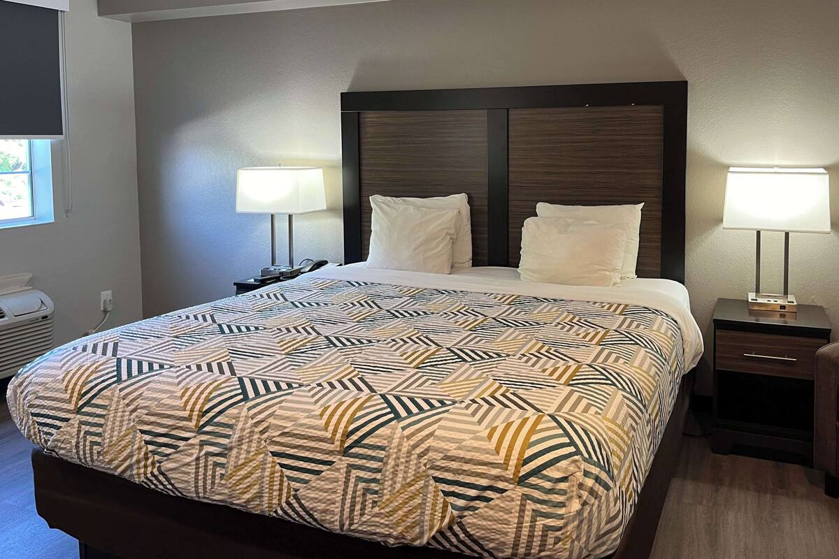 Une chambre d’hôtel avec un grand lit, deux tables de chevet avec des lampes, un radiateur fixé au mur et une fenêtre avec des stores.