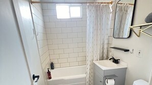 A compact bathroom with white tiled walls, a white bathtub, a white toilet, and a small sink with a mirror.