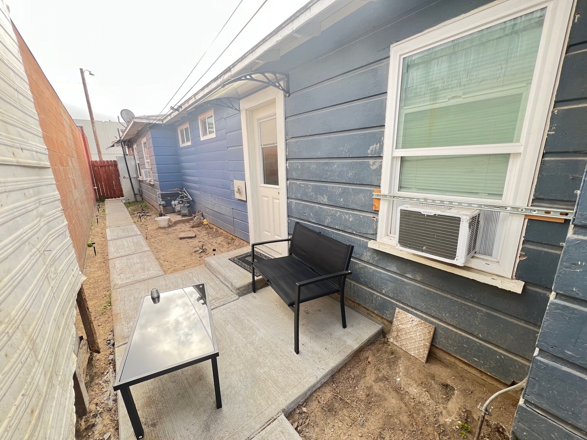 Una casa pequeña de un solo piso con fachada azul, una puerta principal, una ventana con equipo de aire acondicionado, un camino de concreto y un juego de mesa y silla de metal negro.
