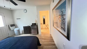 A bedroom with a bed, a ceiling fan, a clock, a trash bin, a small desk with a computer, and a framed picture on the wall.