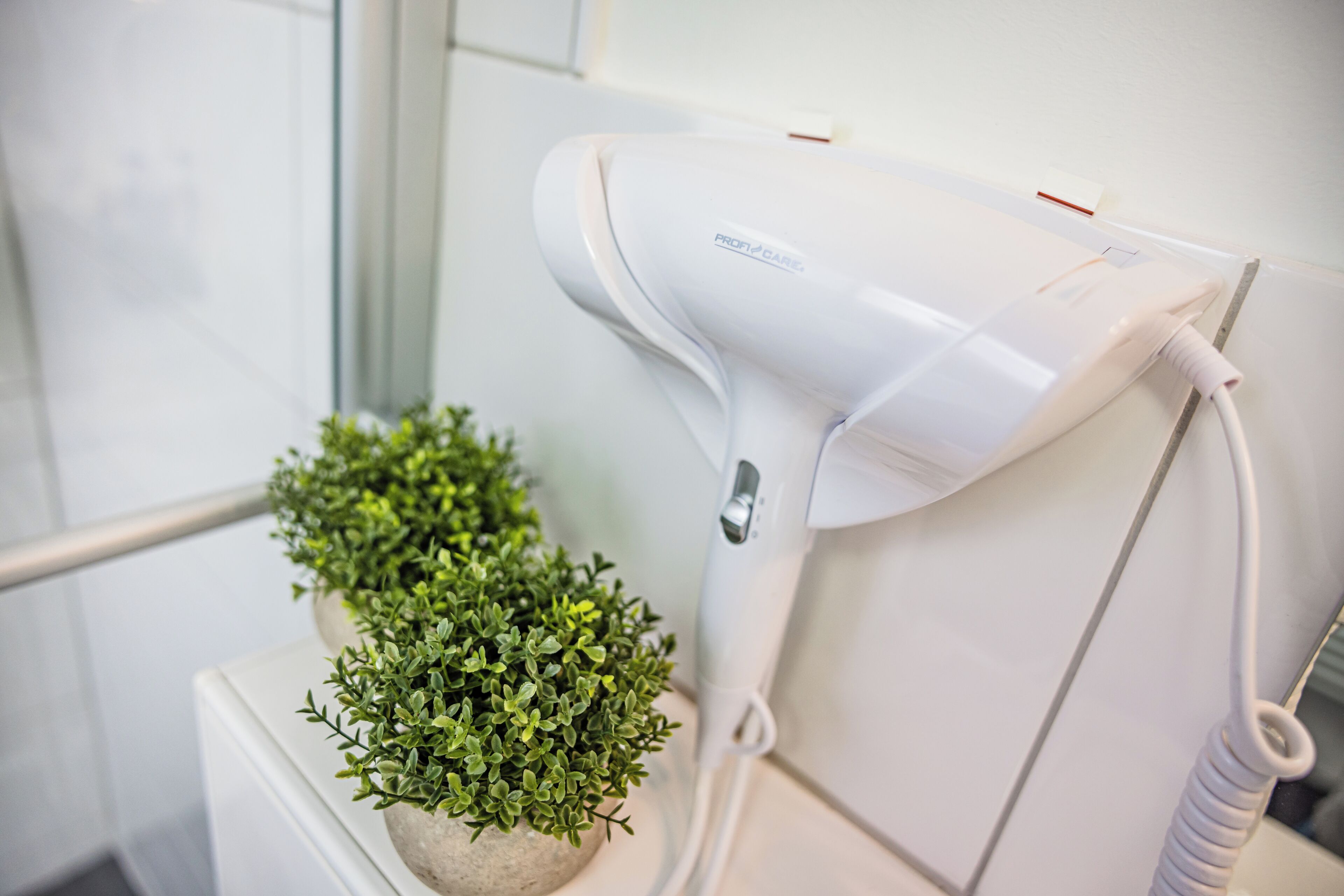 A hair dryer mounted on a white wall, a small potted plant, and a white bathroom counter.