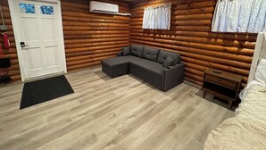Habitación de cabaña de madera con sofá modular, cama, una mesita, ventana con cortinas y equipo de aire acondicionado.