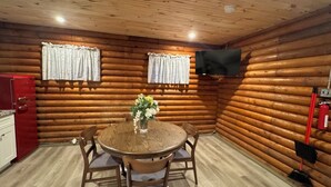 Interior de una cabaña de troncos con una mesa de comedor, sillas, un refrigerador rojo, un televisor montado en la pared y un extintor.