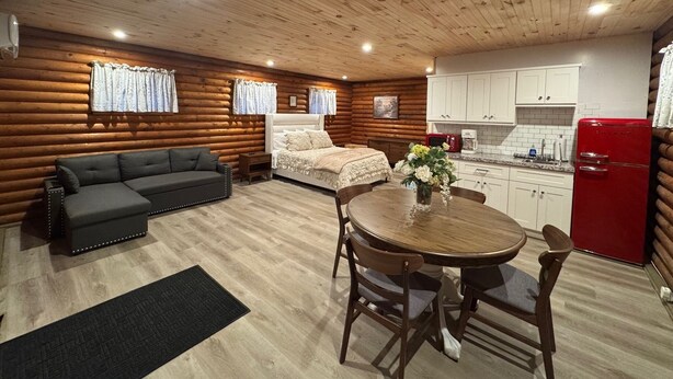 Interior de una cabaña de troncos con sofá, cama, mesa de comedor con sillas, zona de cocina y un refrigerador rojo.