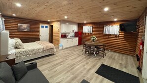 Interior de una cabaña de madera con cama, sofá, mesa de comedor, cocina y televisor montado en la pared.