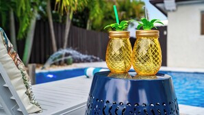 Deux bouteilles en verre doré, dotées de pailles vertes, sont posées sur un présentoir décoratif bleu, près d’une piscine.