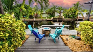 Une terrasse en bois avec deux chaises bleues et un foyer, entourée d’une végétation luxuriante et bordée d’une étendue d’eau paisible.