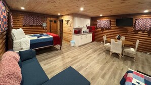 Et hyggeligt værelse i lodge-stil med seng, sofa, spisebord med stole, et tekøkken og et tv ophængt på væggen.