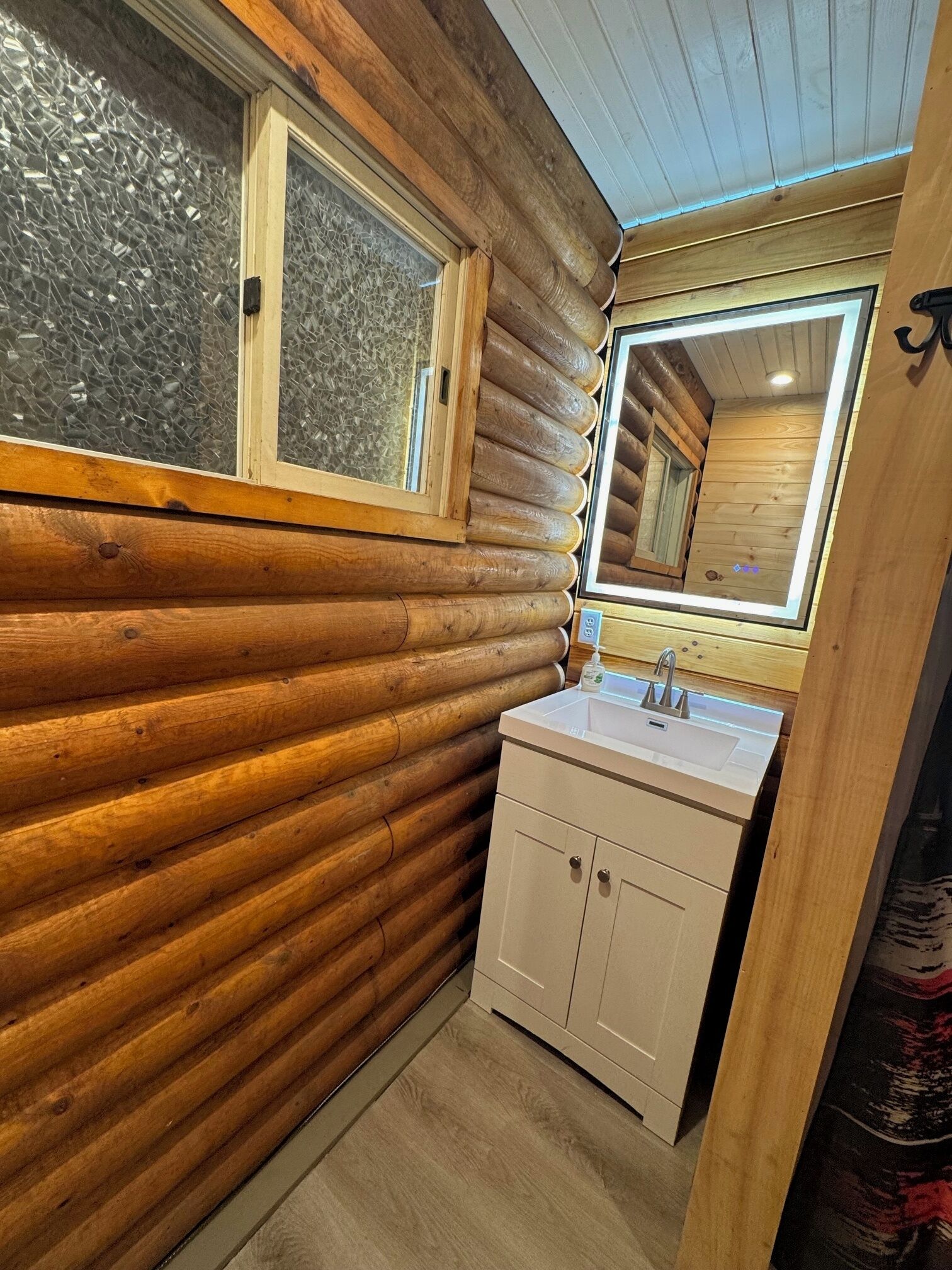 Een houten badkamer in een blokhutstijl, met een buitenkant van hout, een raam met gezandstraald glas, een wastafel met spiegel erboven en een klein kastje.