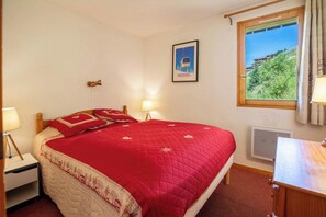 A neatly made bed with a red quilt, a nightstand with a lamp, a framed picture of a ski lift, and a window with a view of greenery and buildings. - Sherpa 11 on the snow front of Mottaret (Les Allues)