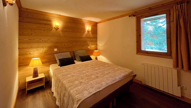 Een slaapkamer met een houten hoofdbord, twee bedlampjes, een bed met beddengoed met een patroon, een nachtkastje en een raam met gordijnen.