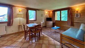 A cozy room with a wooden floor, a dining table with chairs, a small TV, a sofa, and two windows with curtains.