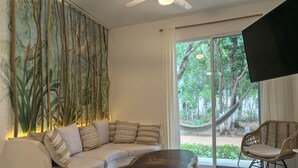 A modern living room with a white sofa, patterned cushions, a wooden coffee table, and a large window with a view of greenery. - 2be 2ba, Playa del Carmen. Ground floor. Free early check in and late check out. (Playa del Carmen)
