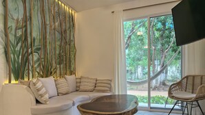 A modern living room with a white sofa, patterned cushions, a round wooden coffee table, a wicker chair, and a large window with a view of greenery. - 2be 2ba, Playa del Carmen. Ground floor. Free early check in and late check out. (Playa del Carmen)