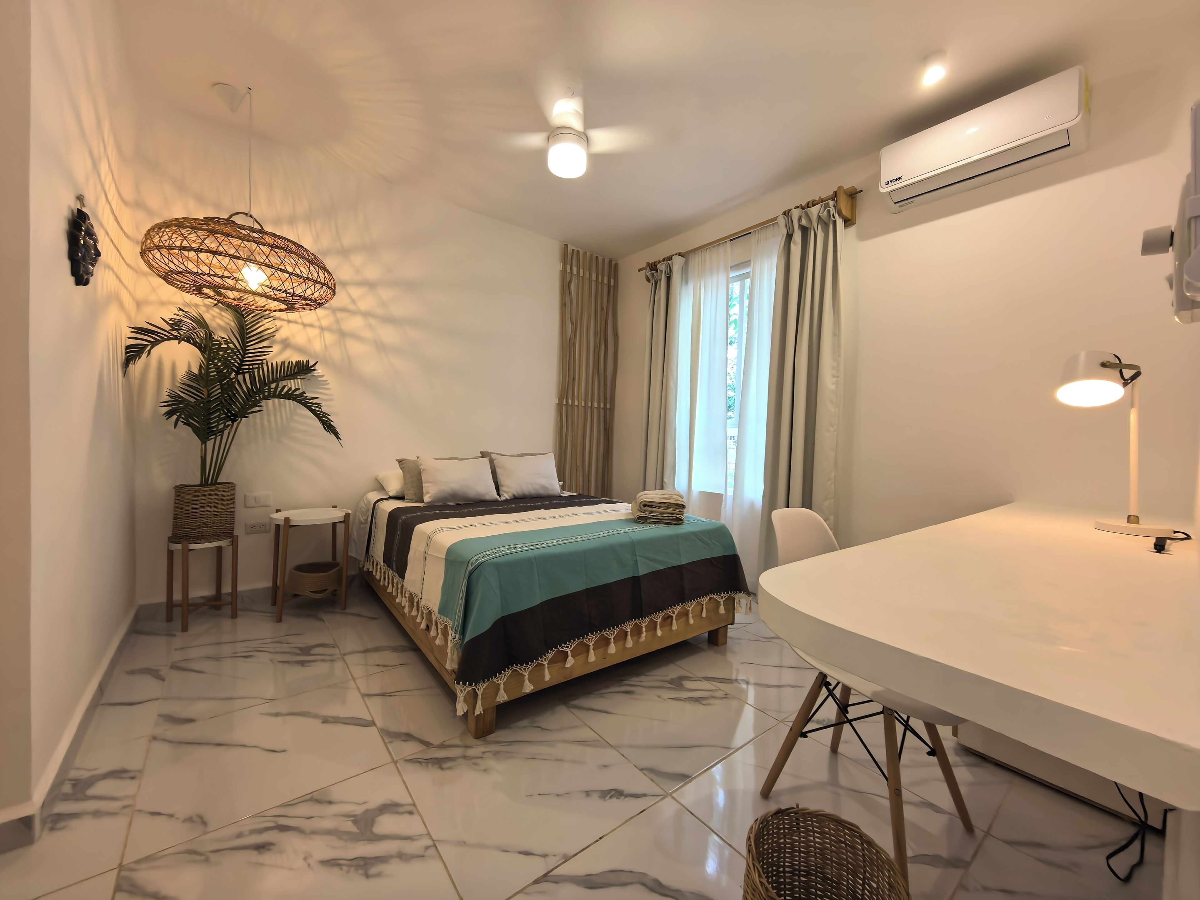 A hotel room with a bed, bedside tables, a desk, a chair, a large potted plant, and an air conditioning unit.