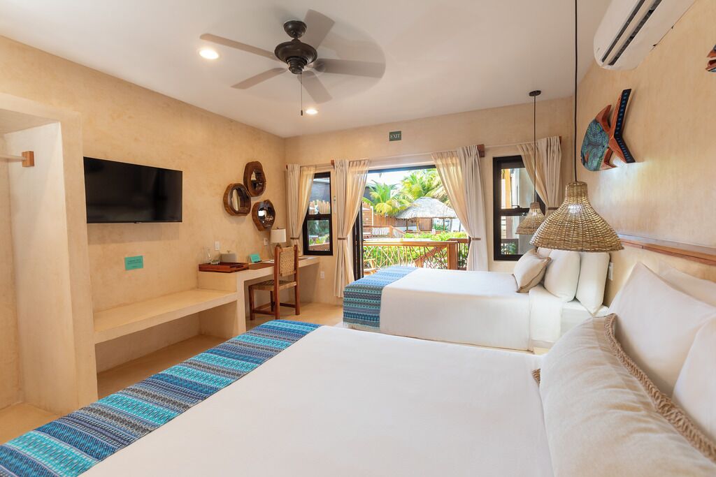 A hotel room with two beds, a desk, a TV, and a balcony with a view of palm trees.