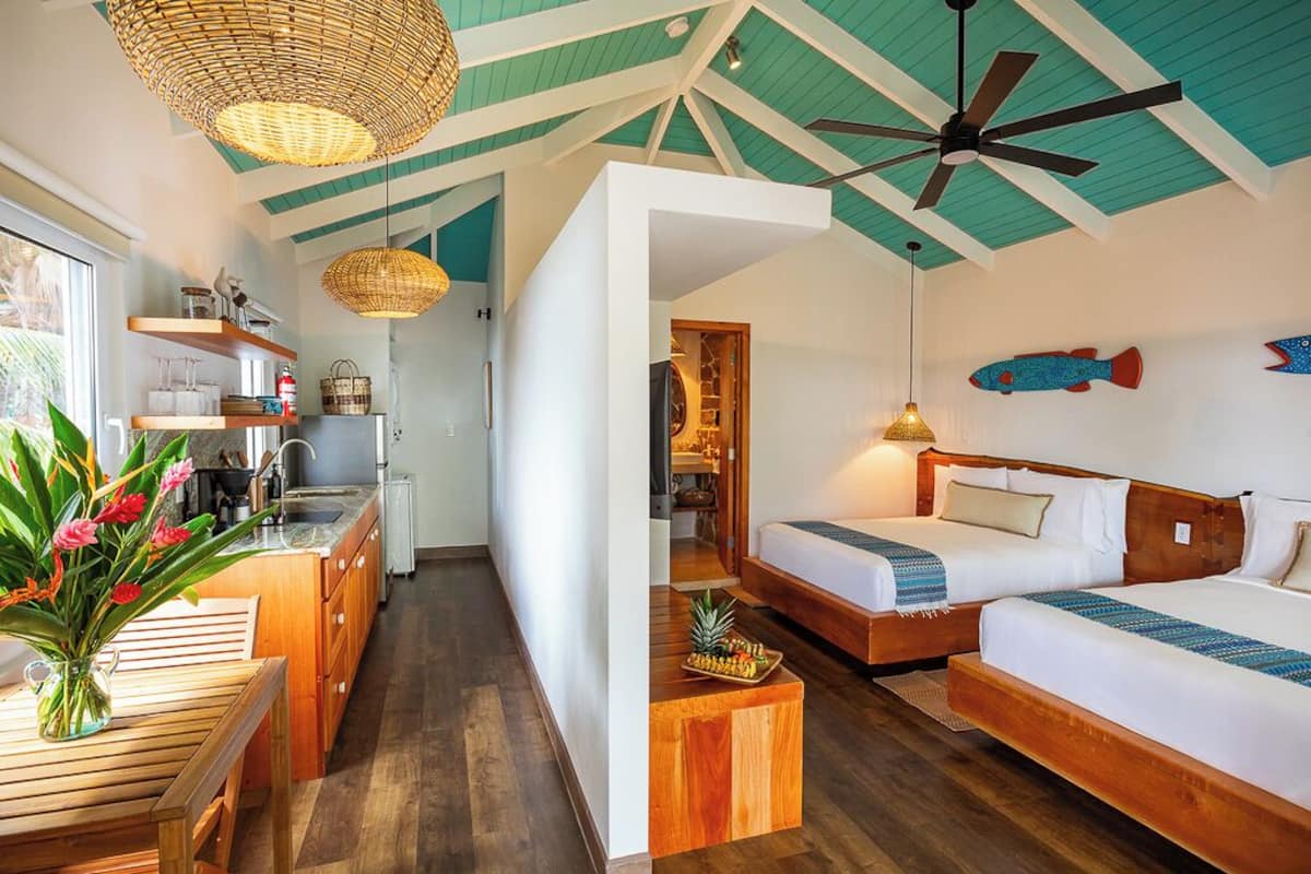 A modern hotel room with a wooden floor, a kitchenette, two beds, and a ceiling fan.
