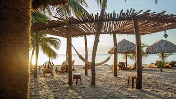 Una playa con sombrillas de paja, sillas de madera y hamacas bajo una pérgola de madera.