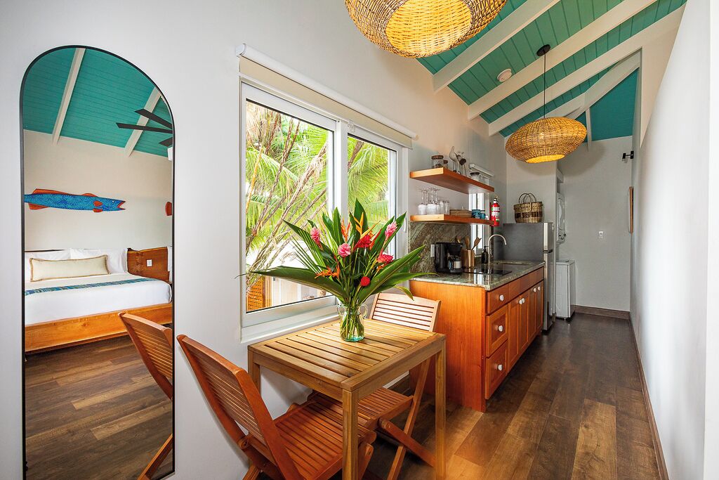 Une pièce compacte comprenant un lit, une table à manger en bois, des chaises, un vase avec des fleurs, un coin cuisine avec une cuisinière et une grande fenêtre donnant sur un paysage verdoyant.