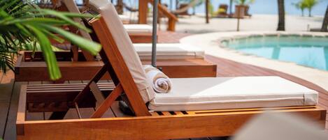 A wooden lounge chair with a rolled towel, a thatched umbrella, and a poolside view.