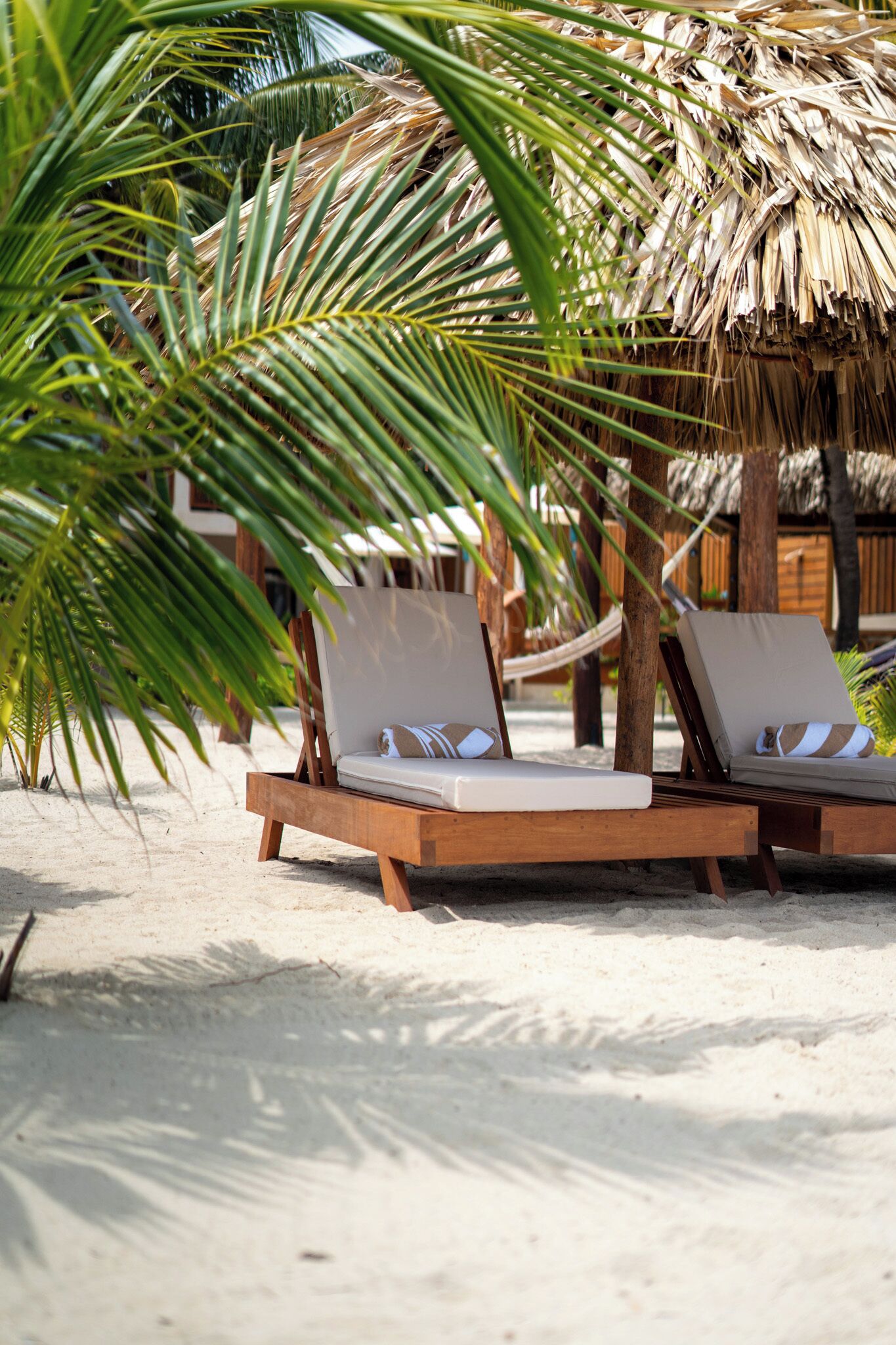 Two wooden lounge chairs with white cushions and striped pillows, positioned under a thatched roof structure on a sandy beach.