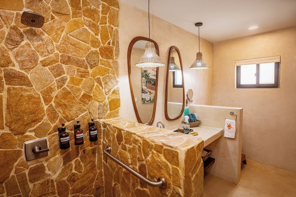 A bathroom with stone walls, a shower, a sink with a mirror, and a window.