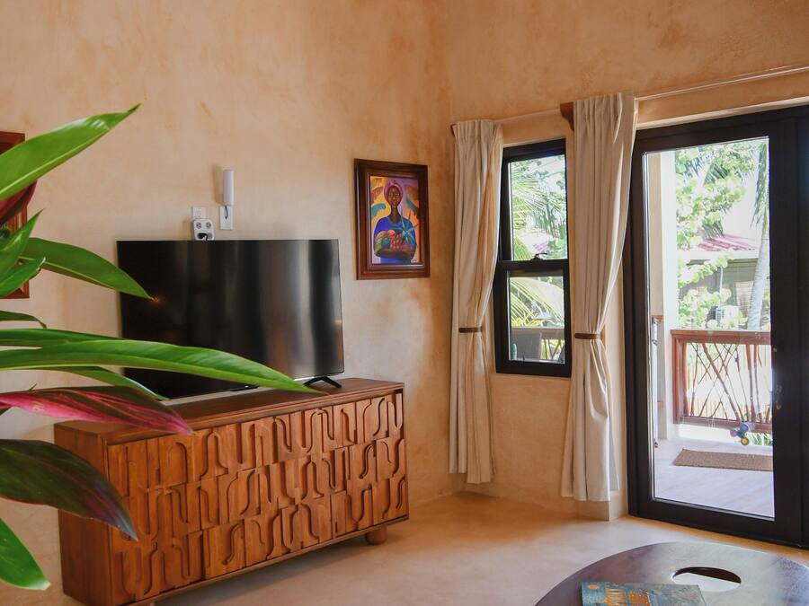 Quarto com televisão sobre um armário de madeira, uma pequena mesa com um livro, uma planta grande e vista pela janela com cortinas.
