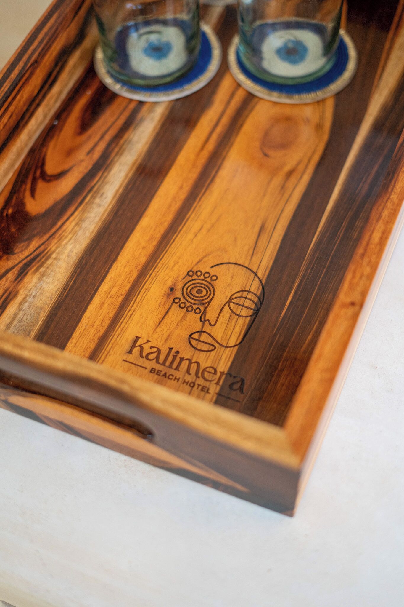 Ein hölzernes Tablett mit zwei Gläsern und einem Logo mit der Aufschrift 'Kalimera Beach Hotel'.
