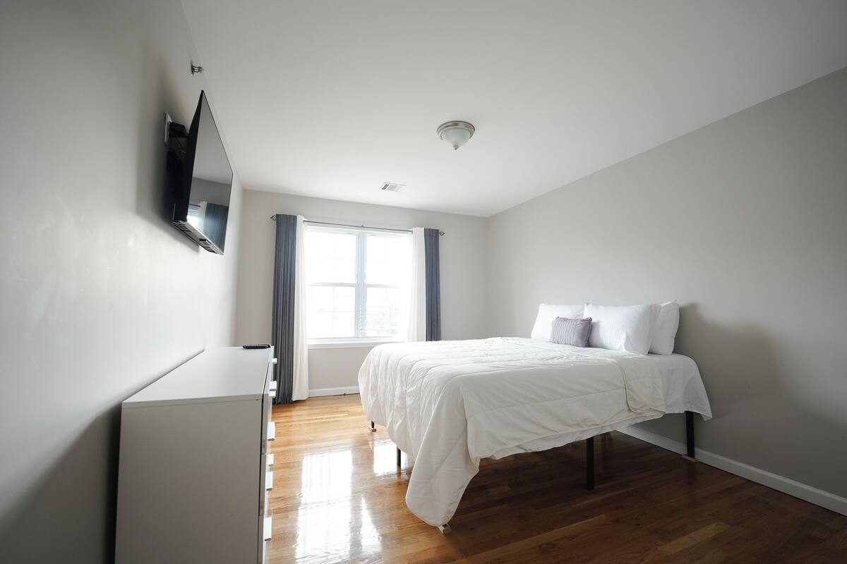Ein Schlafzimmer mit einem Bett, einer Kommode, einem an der Wand montierten Fernseher und einem Fenster mit Vorhängen.