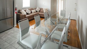 A modern living room with a glass dining table, white chairs, a brown leather sofa, and a kitchen area with a refrigerator.