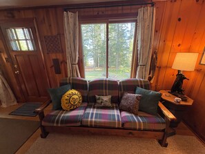 A wooden interior with a plaid sofa, a lamp, and a view of trees through a window.