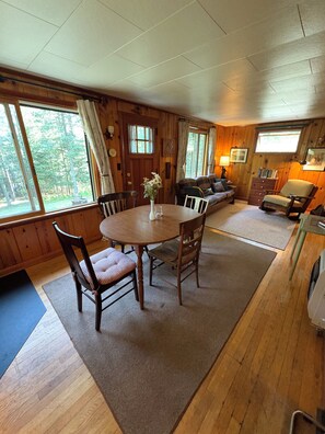 A wooden interior with a dining table, chairs, a sofa, and a window with a view of trees.