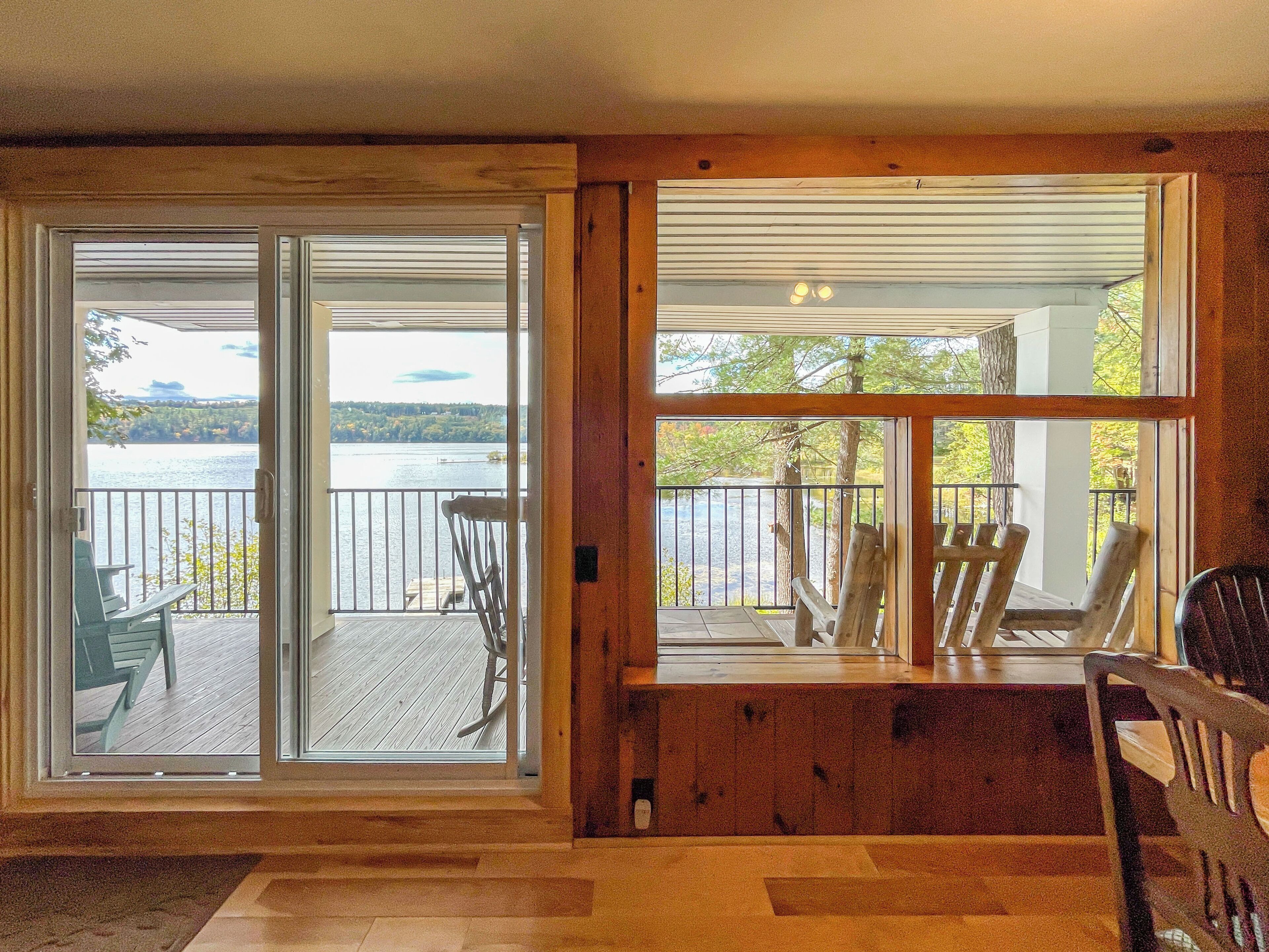 A room with a sliding glass door leading to a balcony with a view of a lake and dock. There are several wooden chairs on the balcony.