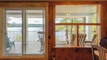 A room with a sliding glass door leading to a balcony with a view of a lake and dock. There are several wooden chairs on the balcony.