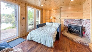 A wooden cabin bedroom with a bed, a fireplace, and a view of a lake through large windows.