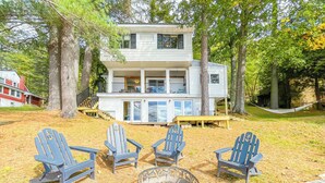 A two-story house with a deck, surrounded by trees. There are four Adirondack chairs and a fire pit in the yard.