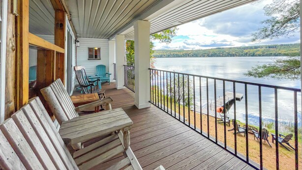 A wooden deck with outdoor seating, overlooking a lake with a view of trees and hills.