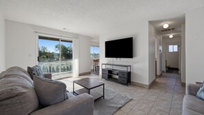 A modern living room with a flat-screen TV mounted on the wall, a sofa, a coffee table, and a sliding glass door leading to a balcony.