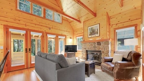 A cozy cabin interior with a stone fireplace, wooden walls, a sofa, a leather armchair, and a television.