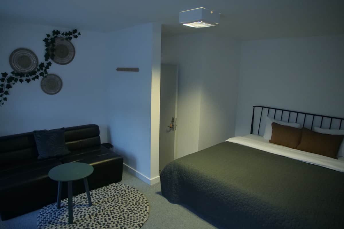 A compact bedroom with a bed, a small sofa, a round table, and a wall-mounted projector.