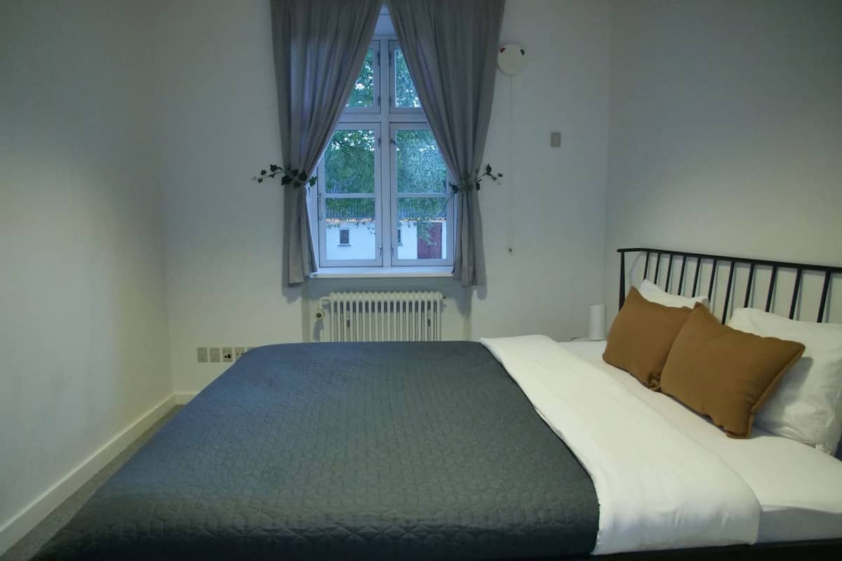 A bedroom with a bed, a window with curtains, a radiator, and a wall-mounted light fixture.