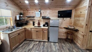 A compact kitchen with wooden cabinets, a refrigerator, a microwave, a sink, and a television mounted on the wall. - Backwoods Bungalow (Pikeville)