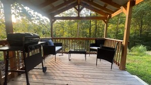 A covered outdoor deck with a grill, two chairs, and a table. - Backwoods Bungalow (Pikeville)