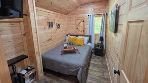 A wooden cabin room with a bed, a small table, a TV, and a window with curtains. - Backwoods Bungalow (Pikeville)