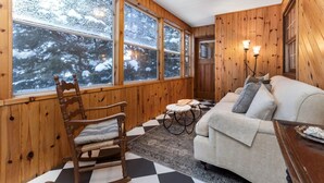 A cozy cabin interior with wooden walls, a checkered floor, a sofa, a rocking chair, and large windows offering a snowy forest view. - 4Bdrm Muskoka Retreat Fire Table, Games, Netflix (Port Carling)