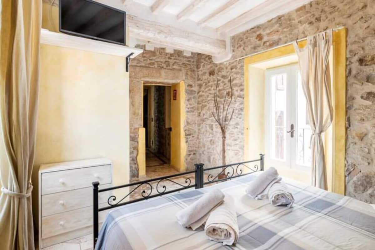 A bedroom with a stone wall, a bed with a plaid bedspread, a bedside table, and a TV mounted on the wall.