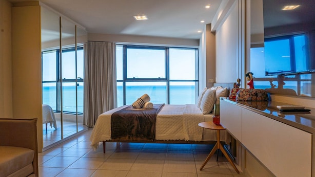 A modern hotel room with a large bed, a small table, a TV, and a view of the sea through large windows. - Flat Beach Class Internacional por Yolo  (Recife)
