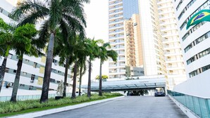 A modern urban street with high-rise buildings, palm trees, and a pedestrian overpass. - Flat Beach Class Internacional por Yolo  (Recife)